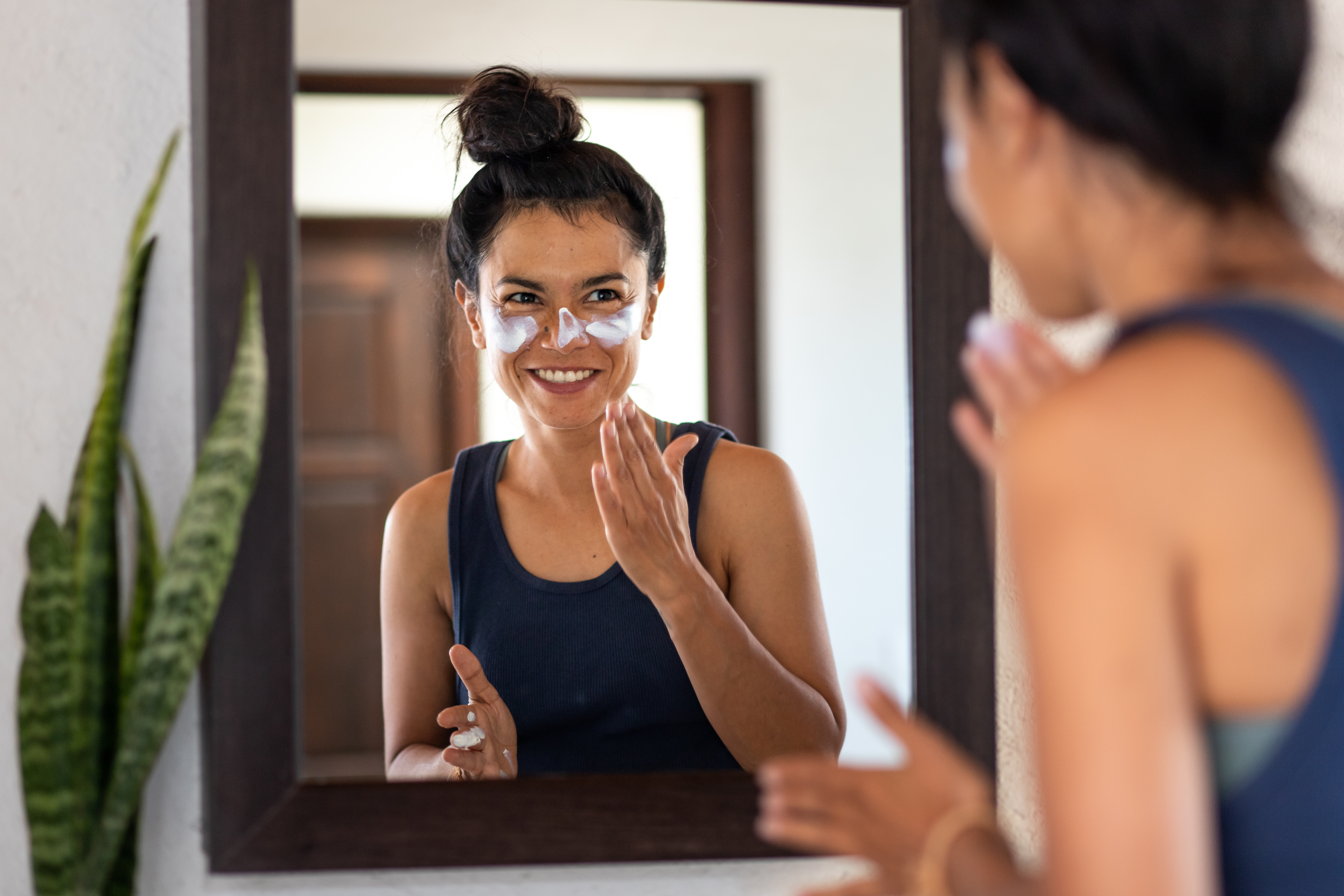 Woman applying white sunblock to face, looking at bathroom mirror, smiling