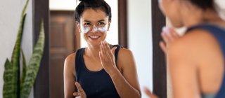 Woman applying white sunblock to face, looking at bathroom mirror, smiling